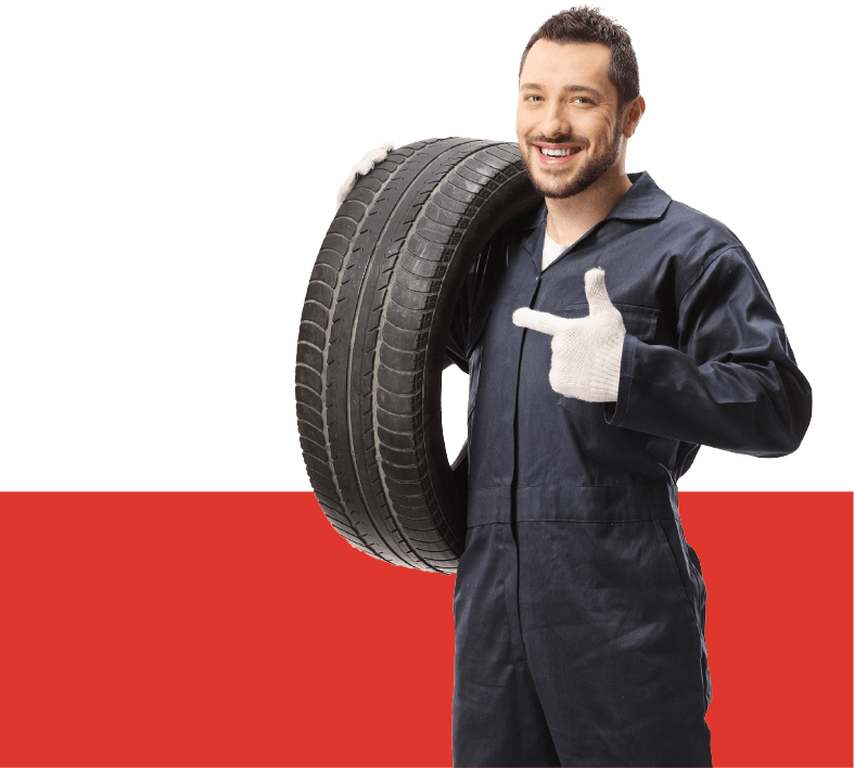 A full shot of an auto mechanic in a gray suit carrying a tire on his shoulder.